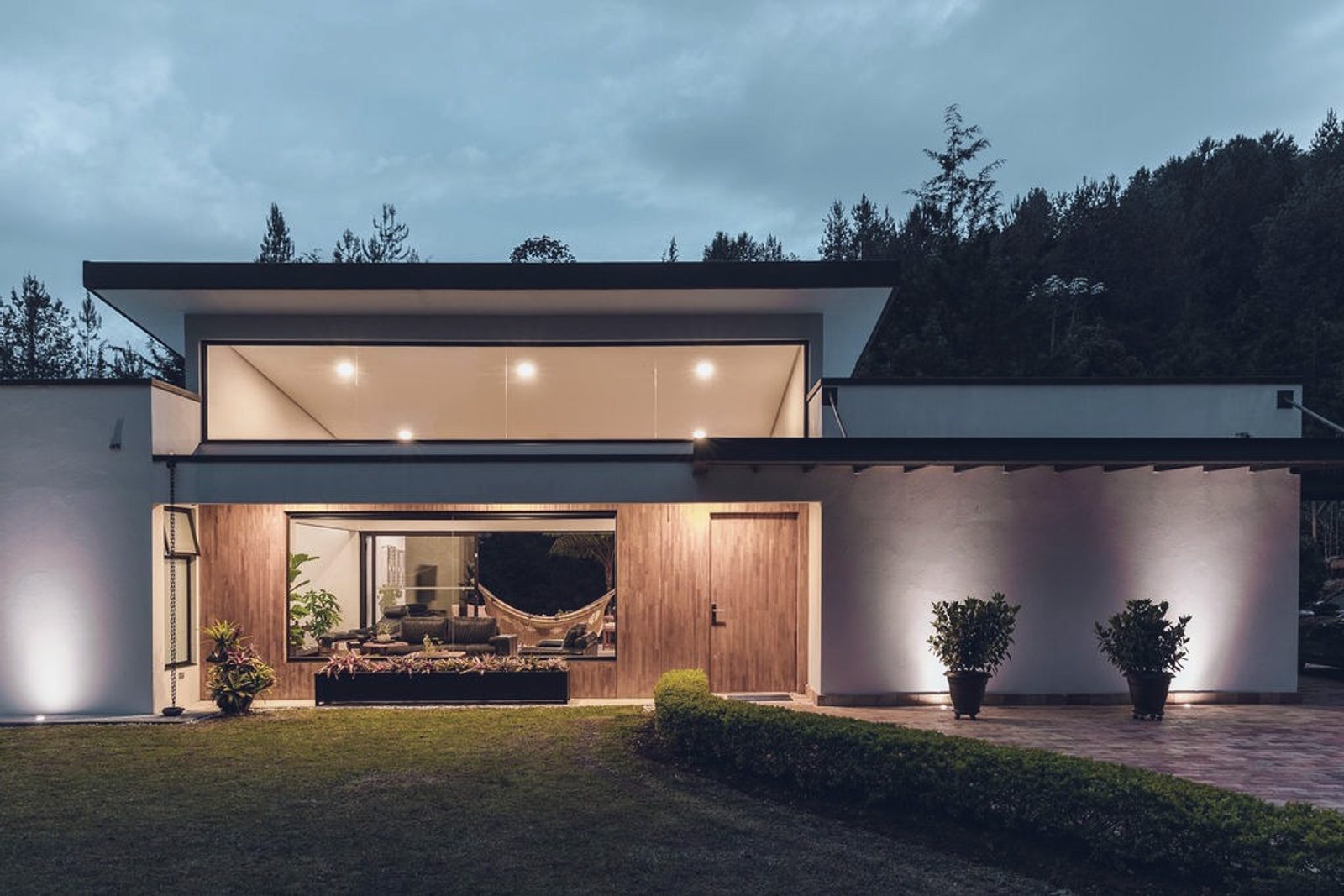 Modern single-story residence at dusk, warm interior light under a broad flat roof against a forested hillside
