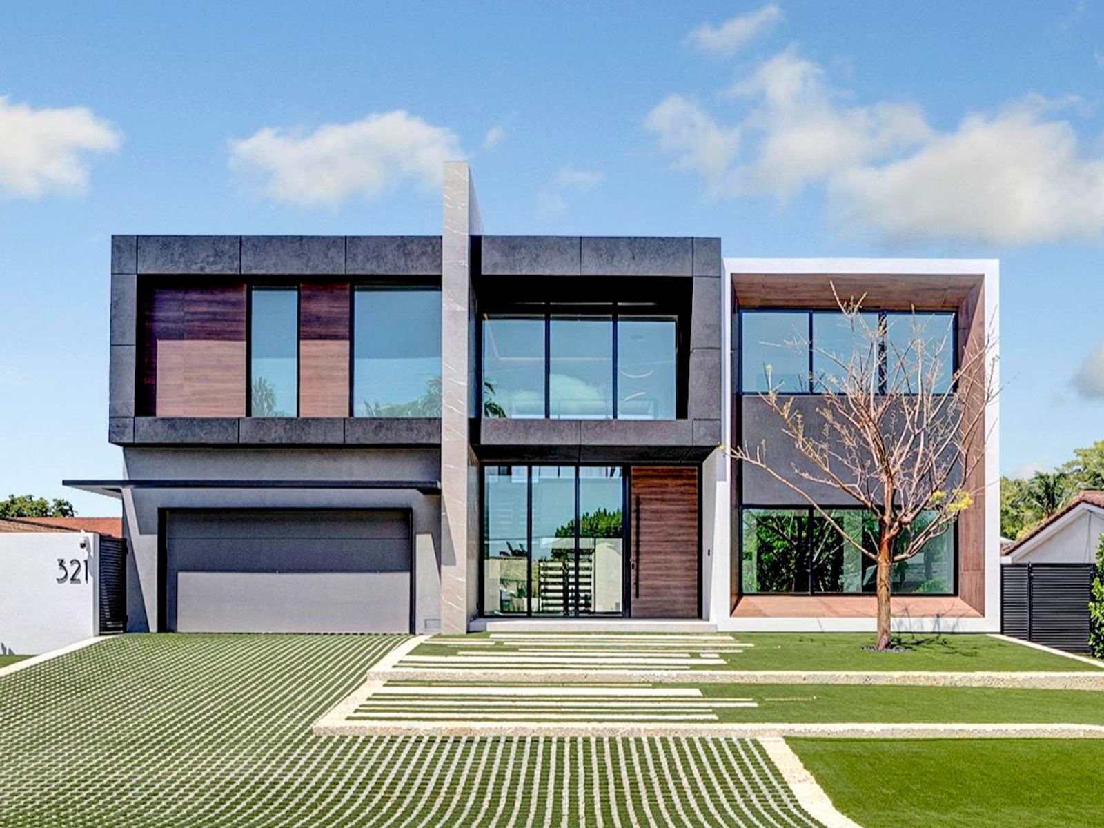 Two-story modern residence with dark stone and wood-clad volumes over a ribbon-grass drive